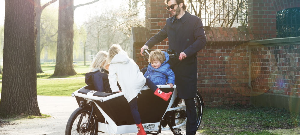 Urban Arrow Family steht im Park, dahinter steht ein Mann, im Lastenkorb sitzen zwei Kinder, ein drittes steigt gerade ein