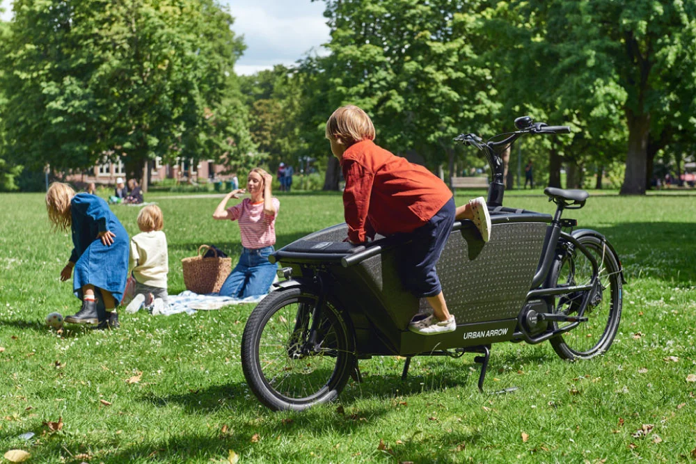 Mutter und Kinder sitzen neben dem Urban Arrow auf der Wiese