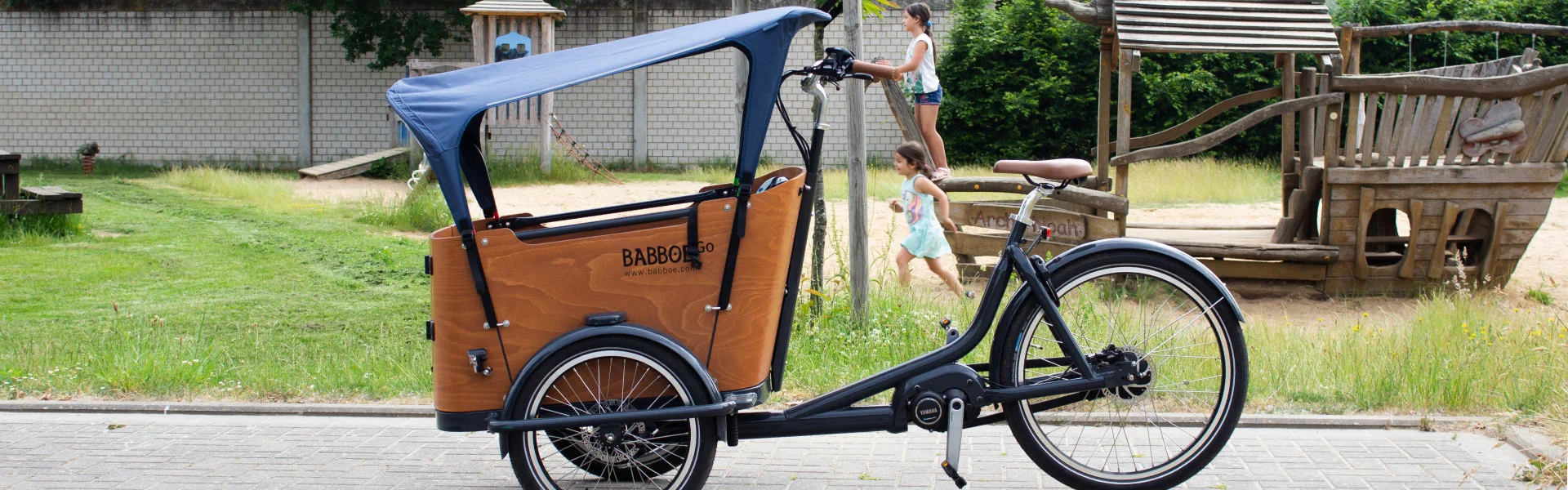 Zwei Kinder spielen auf dem Spielplatz im Vordergrund das Lastenrad Babboe Go Mountain