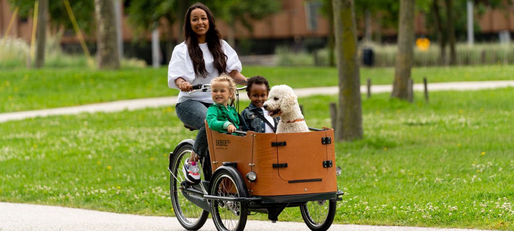 Frau fährt mit Babboe Go Mountain durch einen Park, im Laderaum sitzen zwei Kinder und ein Hund