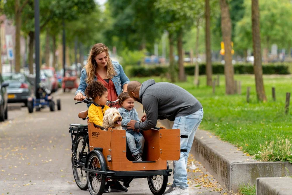Frau steht mit Babboe Go Mountain im Park, im Lastenkorb sitzen zwei Kinder und ein Hund, ein Mann hilft einem Mädchen durch die offene Tür auszusteigen