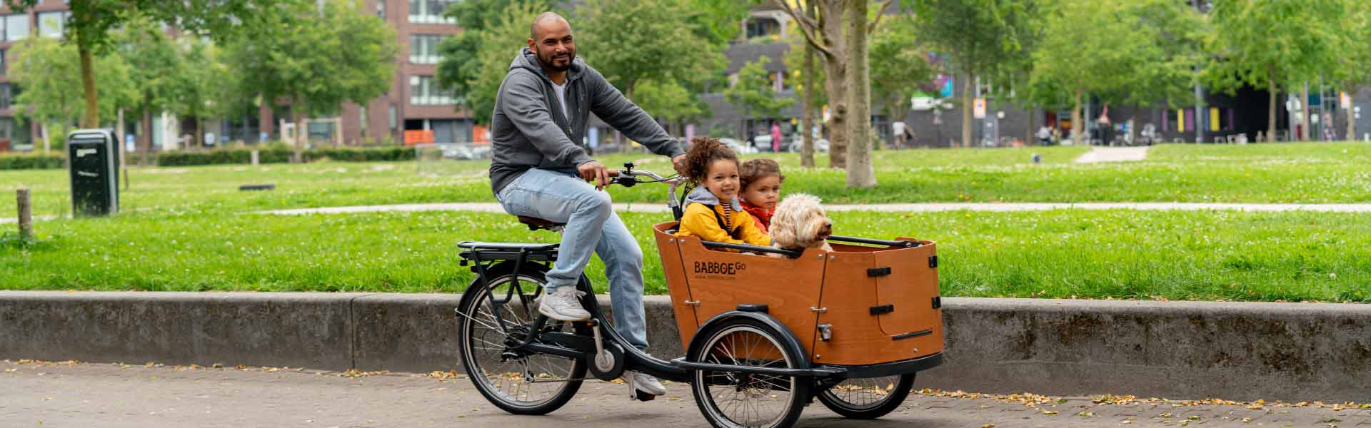 Mann fährt mit zwei Kindern im Laderaum auf dem Babboe Go Mountain durch den Park