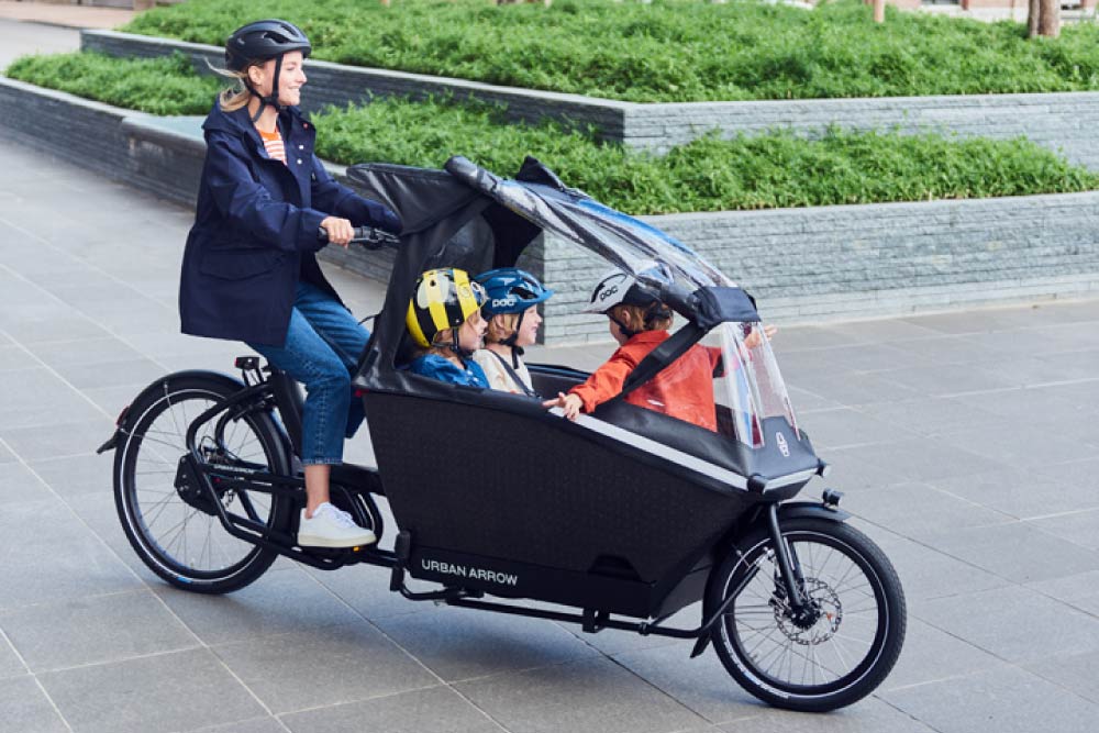 Frau fährt mit Urban Arrow Family durch die Stadt, im Lastenkorb sitzen drei Kinder überdacht von einem Regenschutz