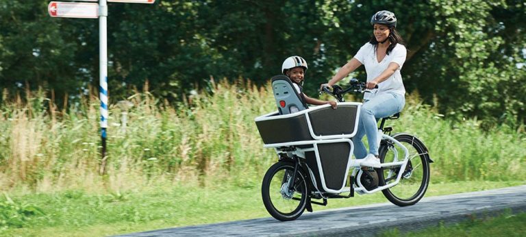 Frau fährt mit dem Urban Arrow Shorty Kids über einen Fahrradweg, im Lastenkorb sitzt ein Kind im Kindersitz